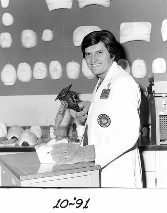 A smiling man in a lab coat holds a tool over a model on a table, with various face molds displayed on the wall behind him.