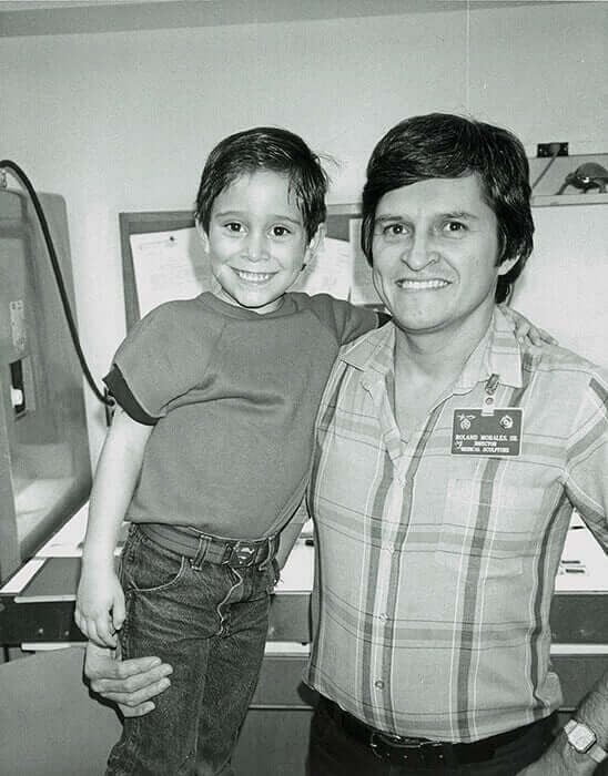 A smiling boy is being held by a man in a plaid shirt. The photo is in black and white, with a casual indoor setting.