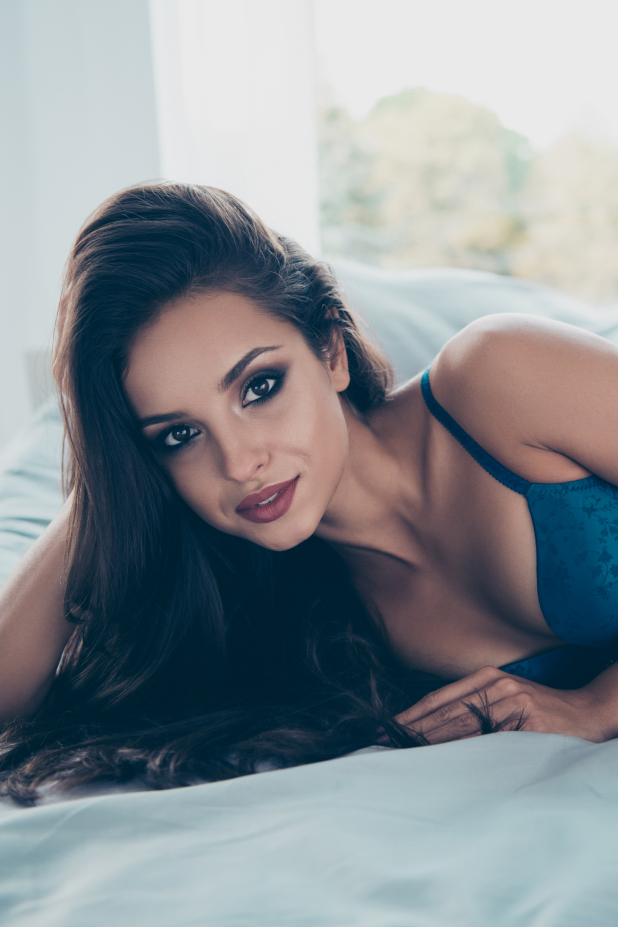 Woman in lingerie laying on bed, head propped up by her hand smiling at camera