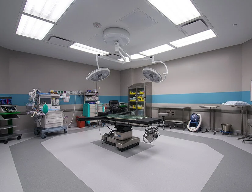 A modern medical room with several hospital beds, privacy curtains, and medical equipment. The space is well-lit and organized.