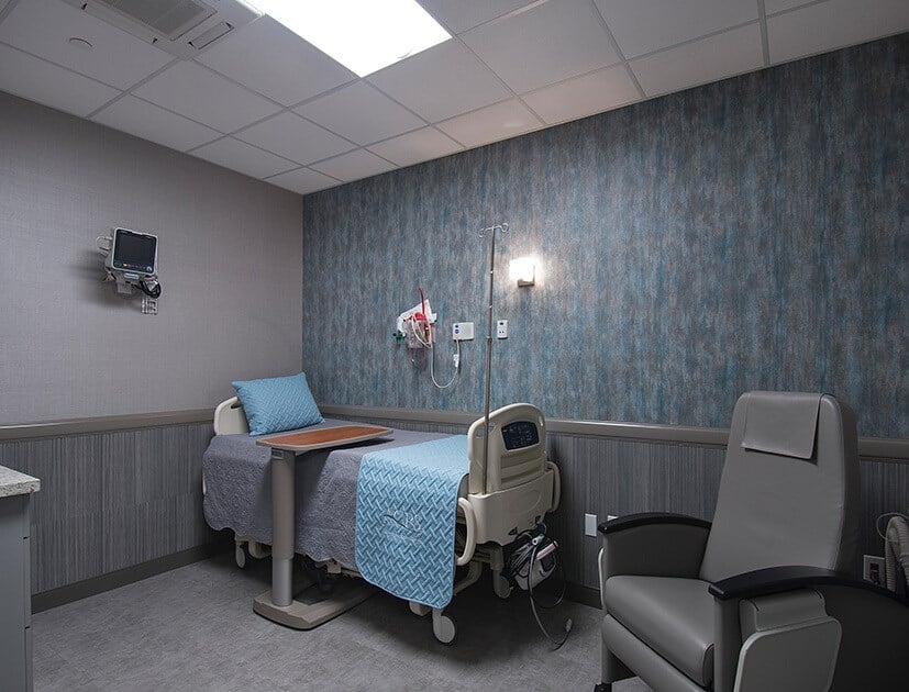 A hospital room featuring a bed with blue bedding, a chair, medical equipment, and a textured blue wall.