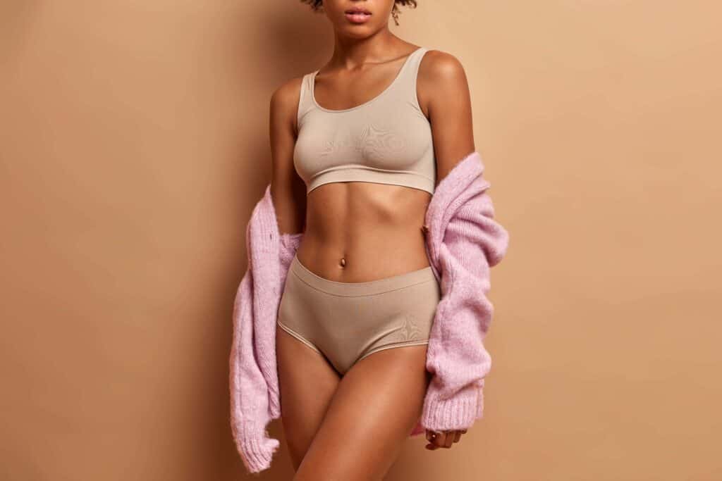 A woman in beige undergarments and a pink cardigan poses against a neutral background, showcasing a toned figure, representing the transformative results of a Mommy Makeover at Dr. Morales' clinic.