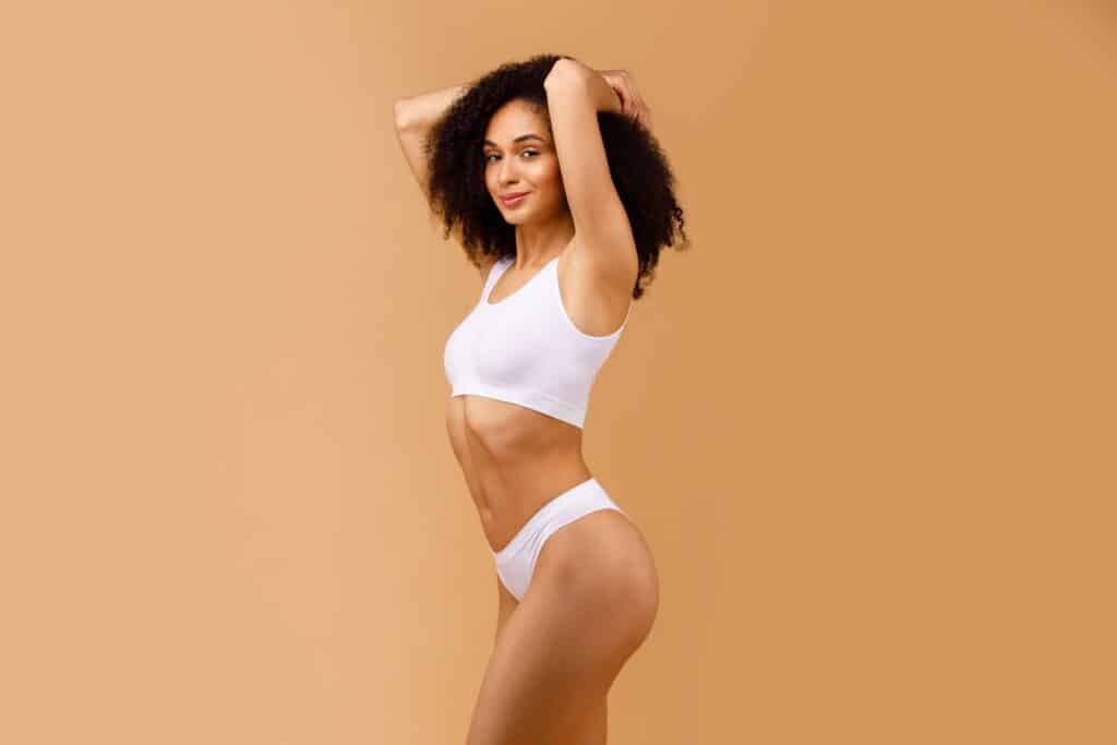 Side view of a fit woman with a toned body and curly hair wearing a white sports bra and underwear, highlighting the transformative results of a Mommy Makeover at Dr. Morales' clinic, emphasizing natural, balanced contours and rejuvenation.