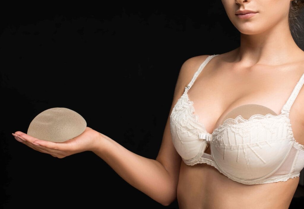 A woman in a white lace bra holds a silicone breast implant in her hand, with another implant visible under her bra, representing the process of breast revision surgery. The breast revision cost varies based on factors such as implant type, surgical complexity, and additional procedures like a breast lift.