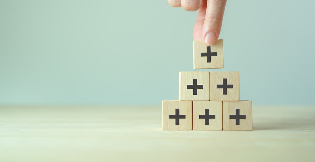 Hand stacking wooden blocks with medical cross symbols in pyramid formation, representing comprehensive healthcare benefits and extended surgical procedure advantages