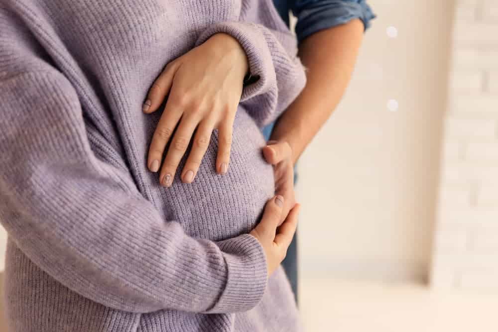 Close-up of a pregnant woman in a cozy sweater holding her belly, representing considerations for a tummy tuck after pregnancy.