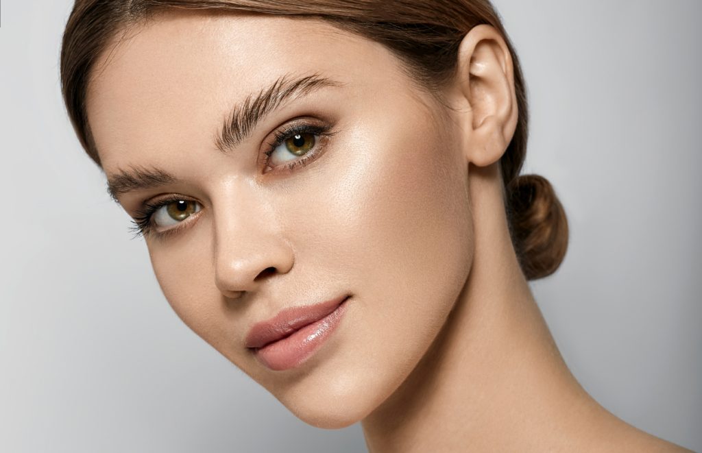 Front-facing close-up of a woman with minimal makeup, hair pulled back, and even facial lighting.