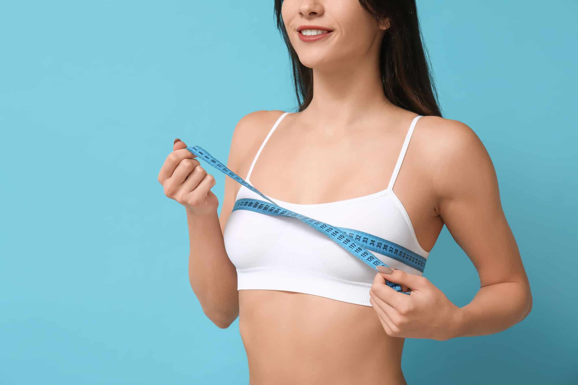 A woman in a white sports bra smiling while measuring her chest with a blue measuring tape, highlighting the benefits of breast reduction.