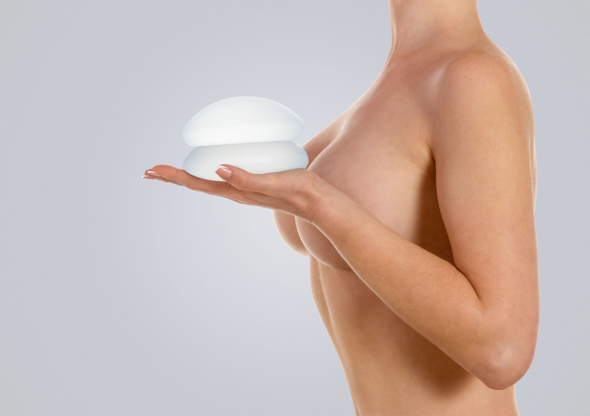 A side profile of a woman holding two silicone breast implants in her hand near her chest to visualize breasts after implant removal.